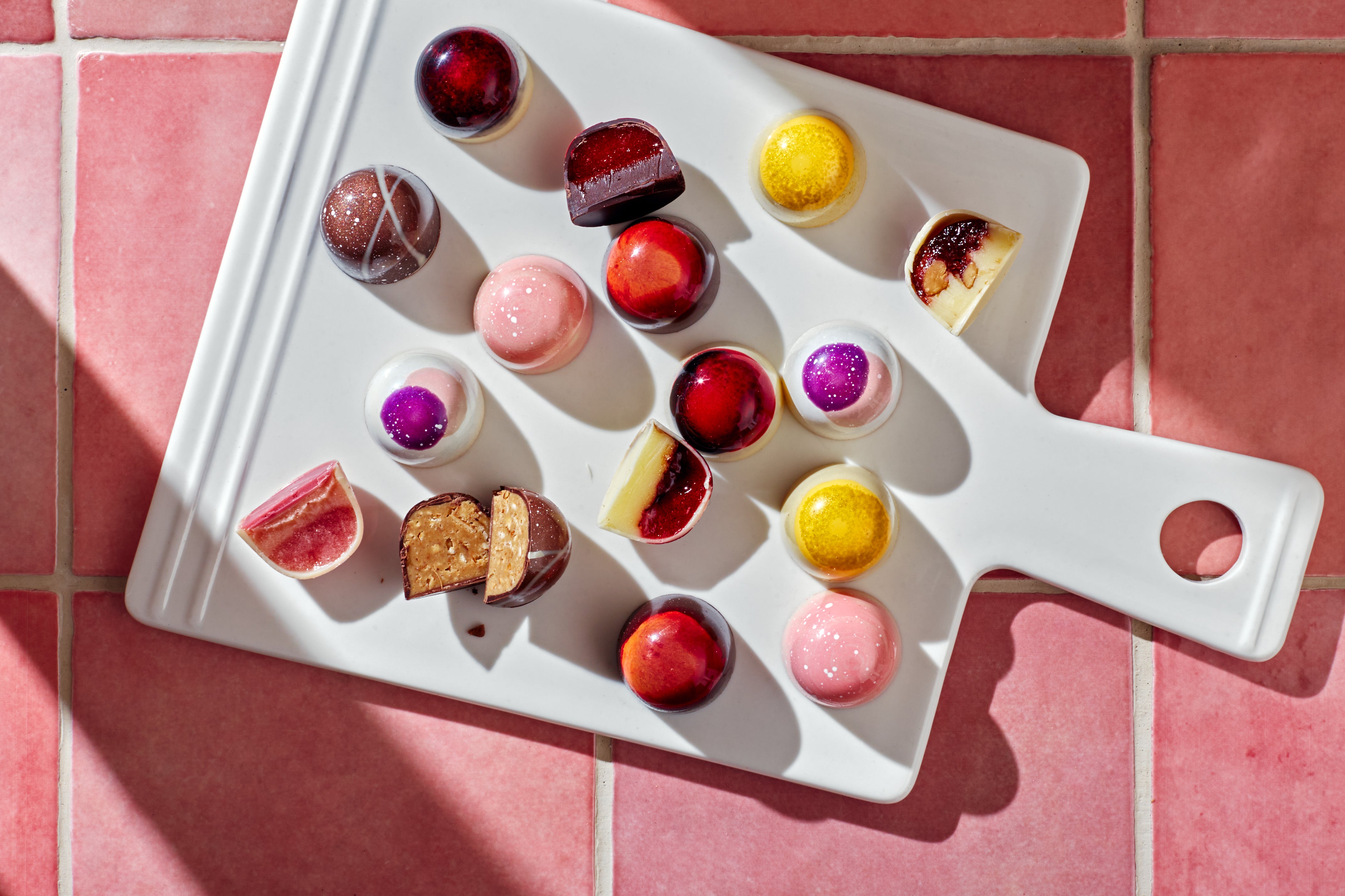 An array of colourful bon bons in spring hues, placed on a white cutting board on a pink tile background. 